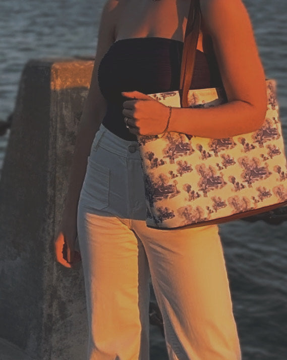 Up close image of a woman in white jeans, a blue strapless bodysuit, holding a large blue and white toile tote bag that features the architecture of Beaufort,SC. The woman is standing in waterfront park in beaufort,sc.