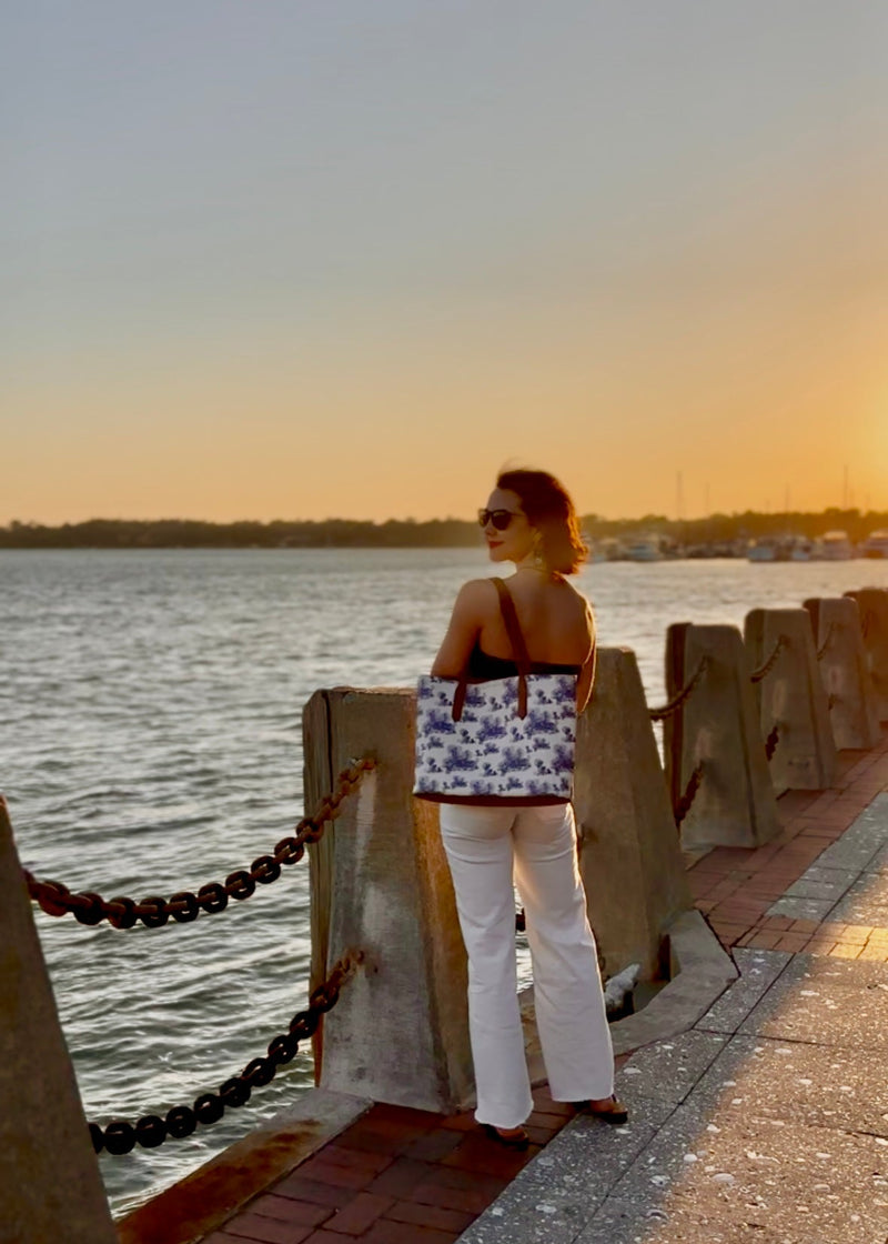 Woman standing by a waterfront at sunset, wearing a navy blue strappless top and white raw-hem jeans. She's carrying a blue and white toile tote bag that features the architecture of Beaufort, SC.